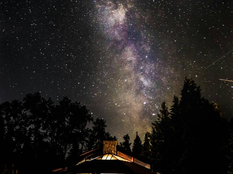 A serene night sky with the Milky Way above a lit cabin, surrounded by trees.